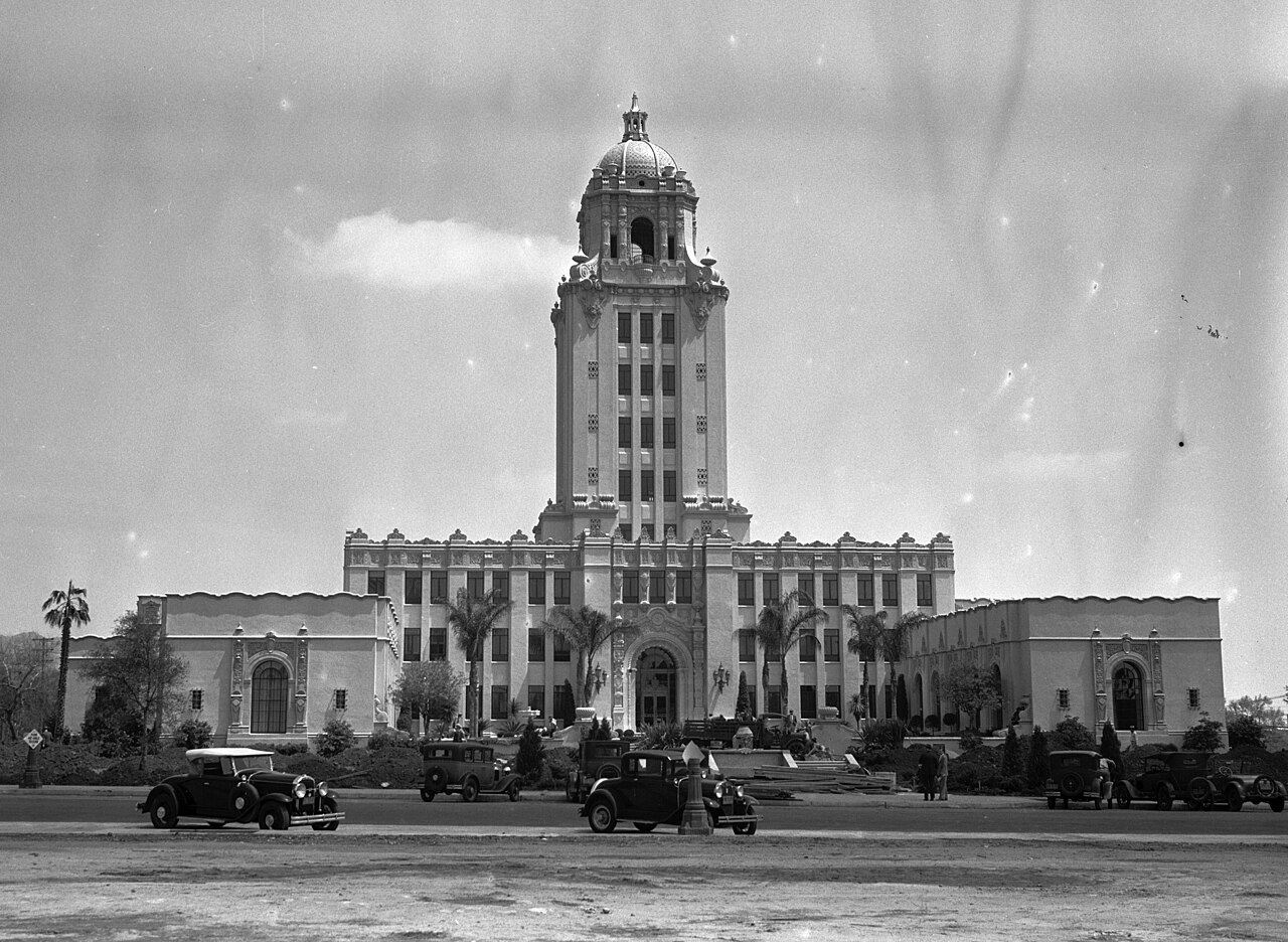 A black-and-white view of a low building with two wings and a tall central tower topped by a dome, with 1930s cars parked in front.