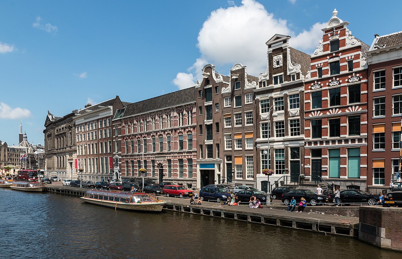 A canal basin with a tour boat moored at the quay in front of tall brick houses with stepped gables under a blue sky.