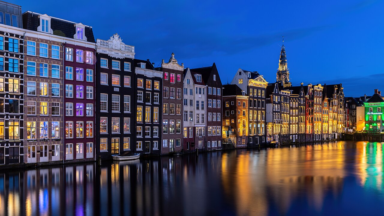 A row of tall narrow gabled houses with lit windows rising straight out of dark water that mirrors their coloured lights.