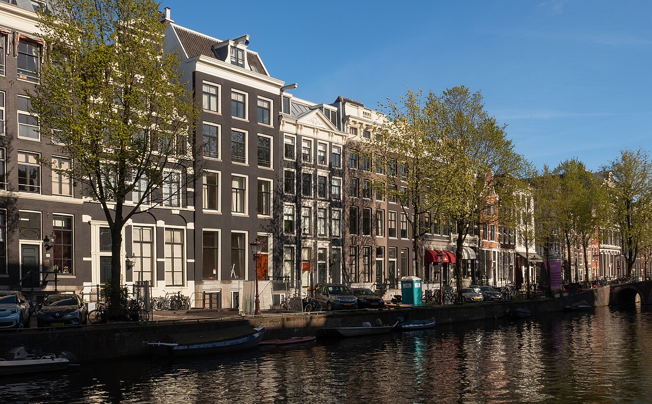 Dark brick canal houses with white-framed windows behind a line of trees, reflected in the still water of a canal.