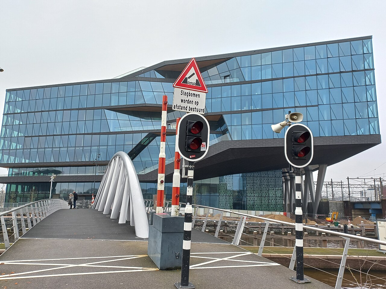 A glass office building with a jutting upper storey behind a footbridge with white arches and red-and-white barrier lights.