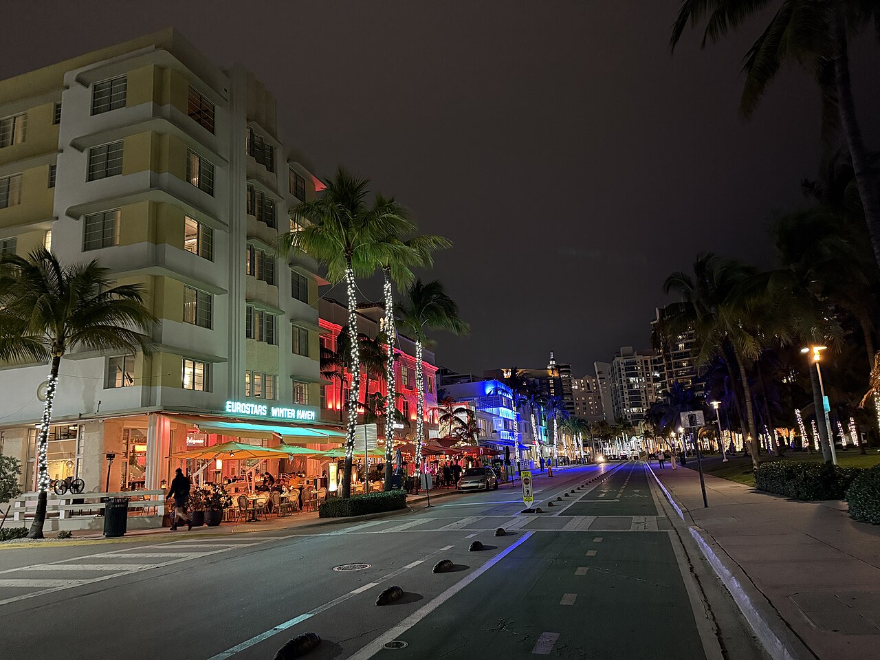 A street at night with palm trees and low hotels lit in neon on the left and an empty road curving away on the right.