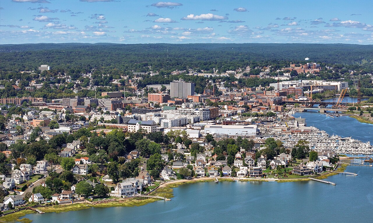 An aerial view of a small city of low buildings and trees around a blue harbour with houses along the shore.