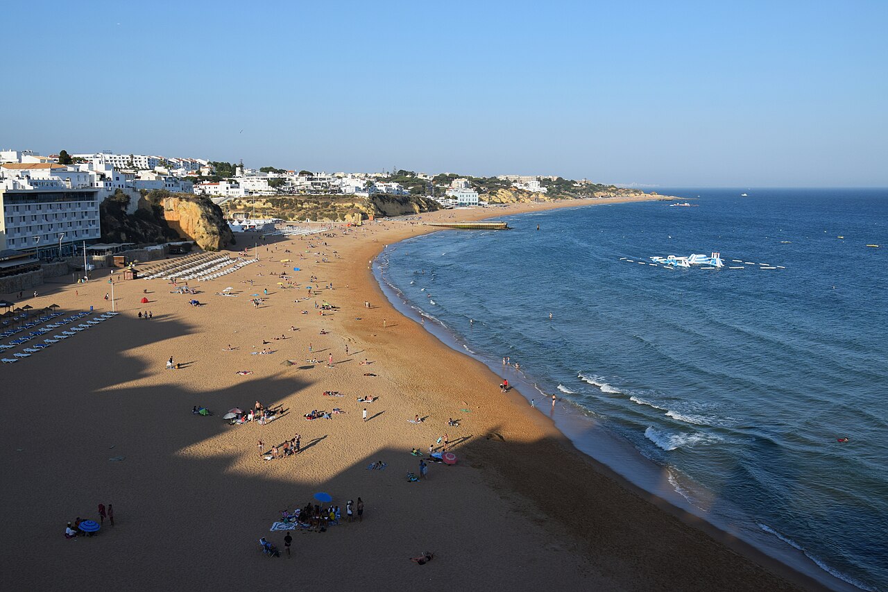 A long curve of golden sand with scattered bathers, blue sea on the right and white buildings on low cliffs behind.