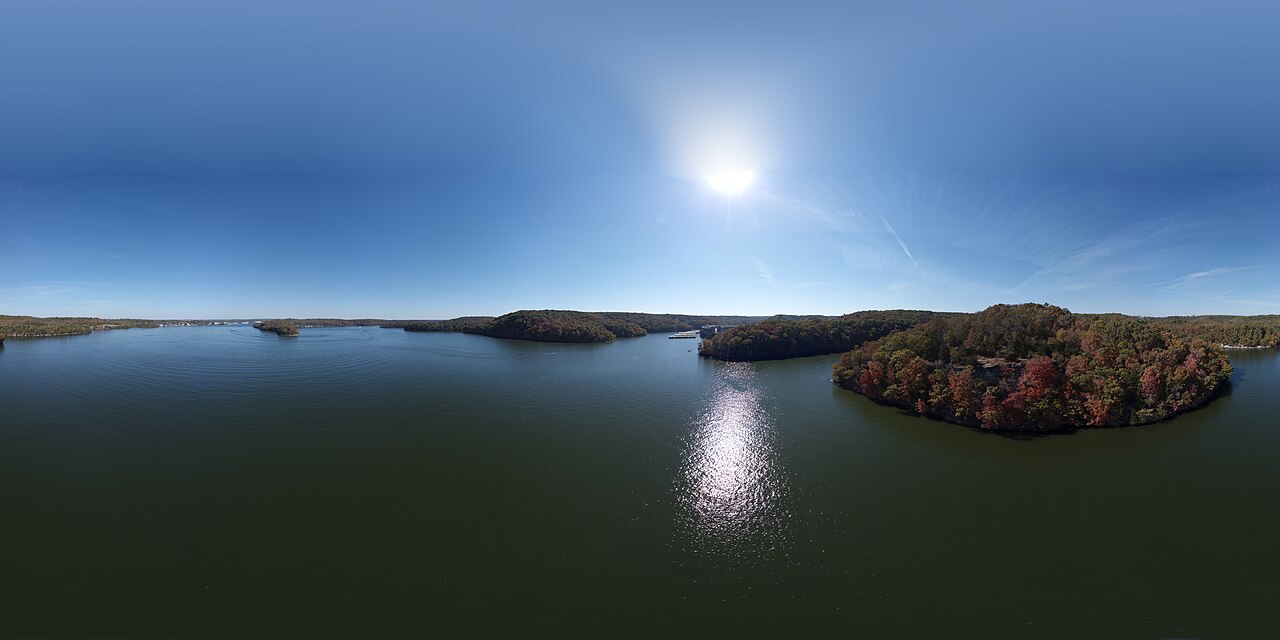 A wide panorama of a dark blue lake winding between wooded shores in autumn colour, with the sun high in a clear sky.