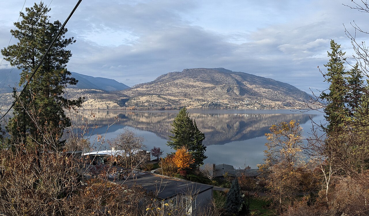 A calm lake reflecting a bare rounded mountain, framed by pine trees and autumn shrubs on a hillside.