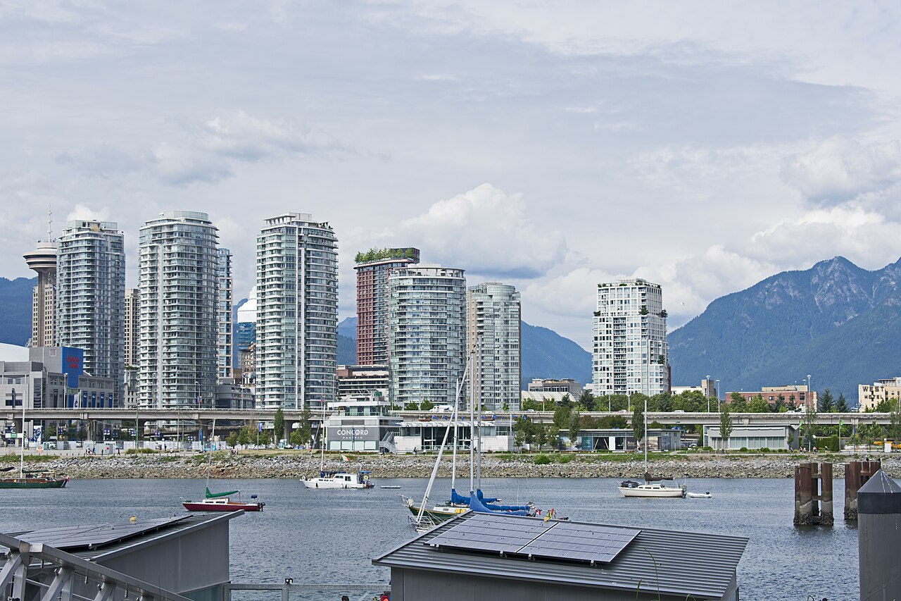 A row of glass residential towers along a waterfront with moored boats in front and mountains behind, under a cloudy sky.