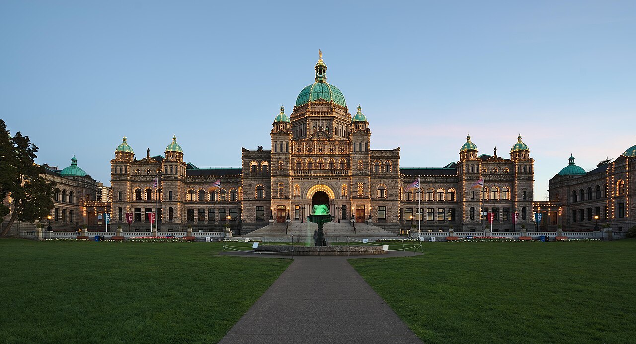 A long stone building with a central green dome, its outline and windows lit, at the end of a path across a dark lawn.