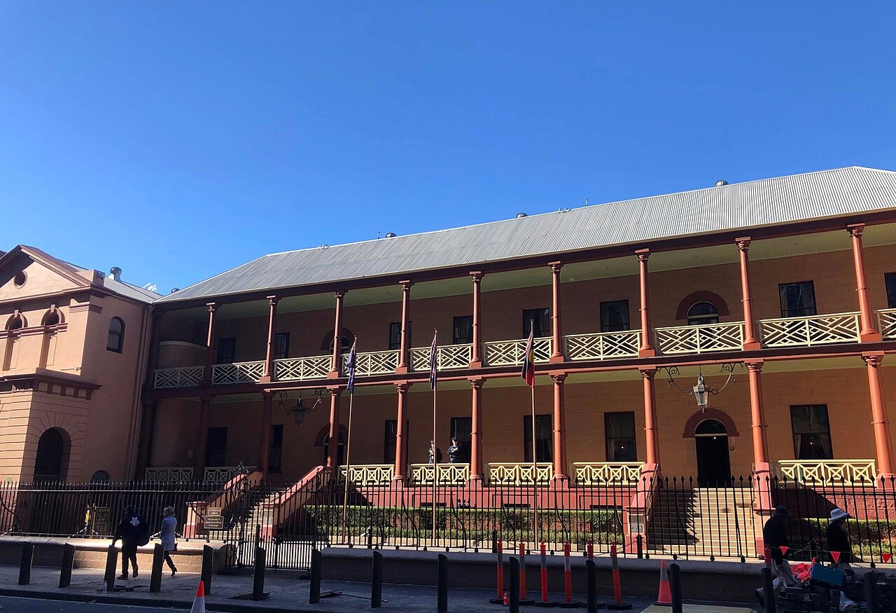 A two-storey colonial building with long verandahs and iron railings in afternoon sun, behind a fence on a city street.