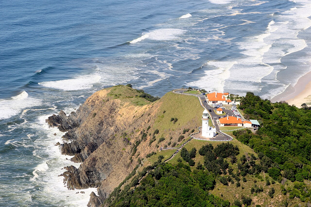 A white lighthouse and red-roofed cottages on a green, rocky headland with lines of white surf breaking on both sides.