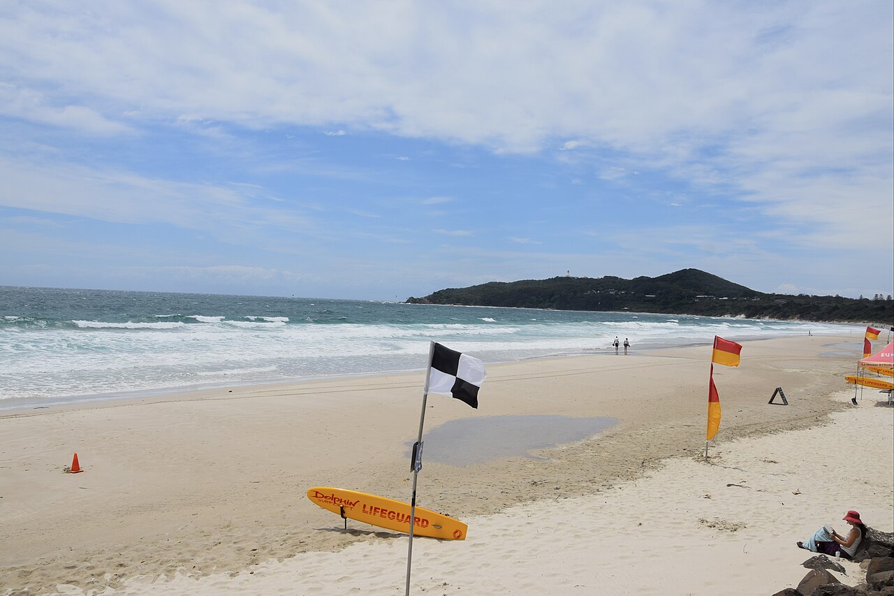 A wide sandy beach with surf lifesaving flags and a rescue board, waves rolling in and a dark headland on the horizon.