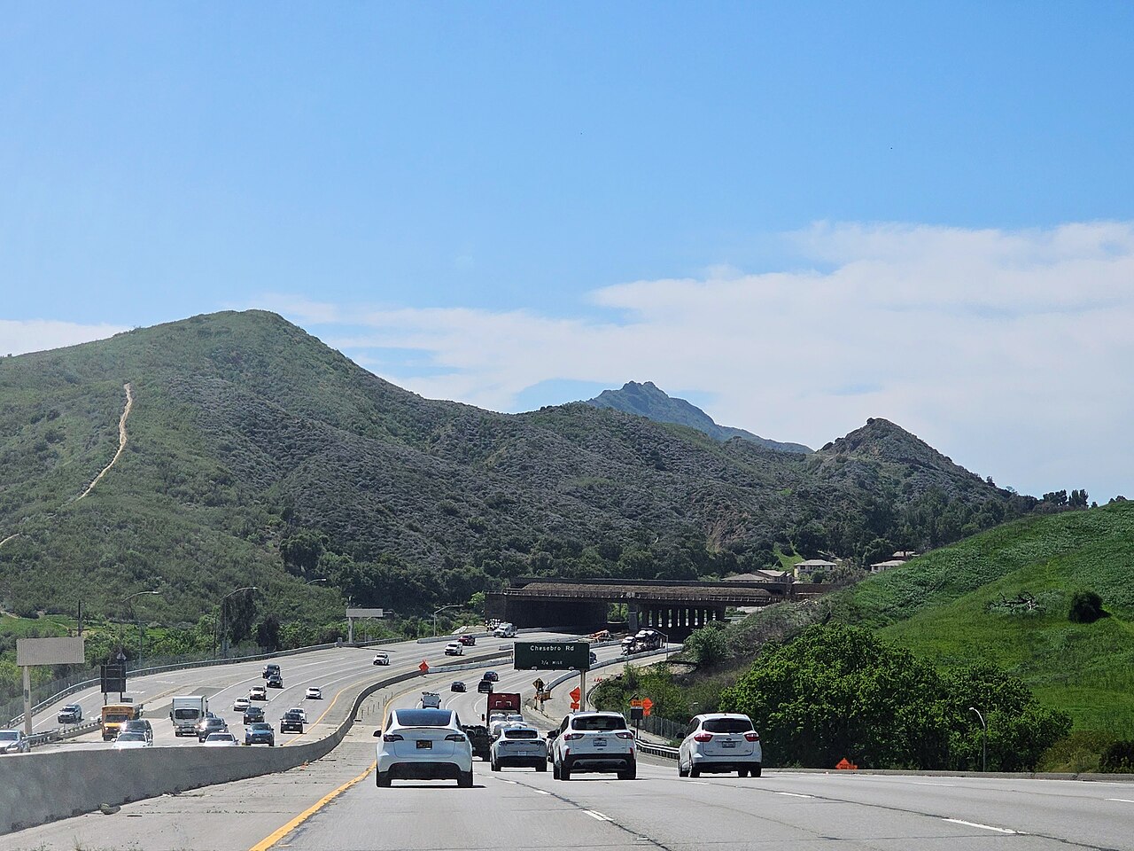 Green hills and rocky peaks under a blue sky, with a busy highway running through the valley at the bottom of the frame.