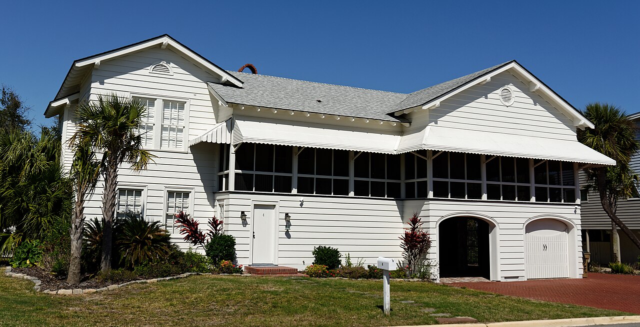 A white wooden house raised a full storey above the ground, with a screened porch, striped awnings and palm trees.