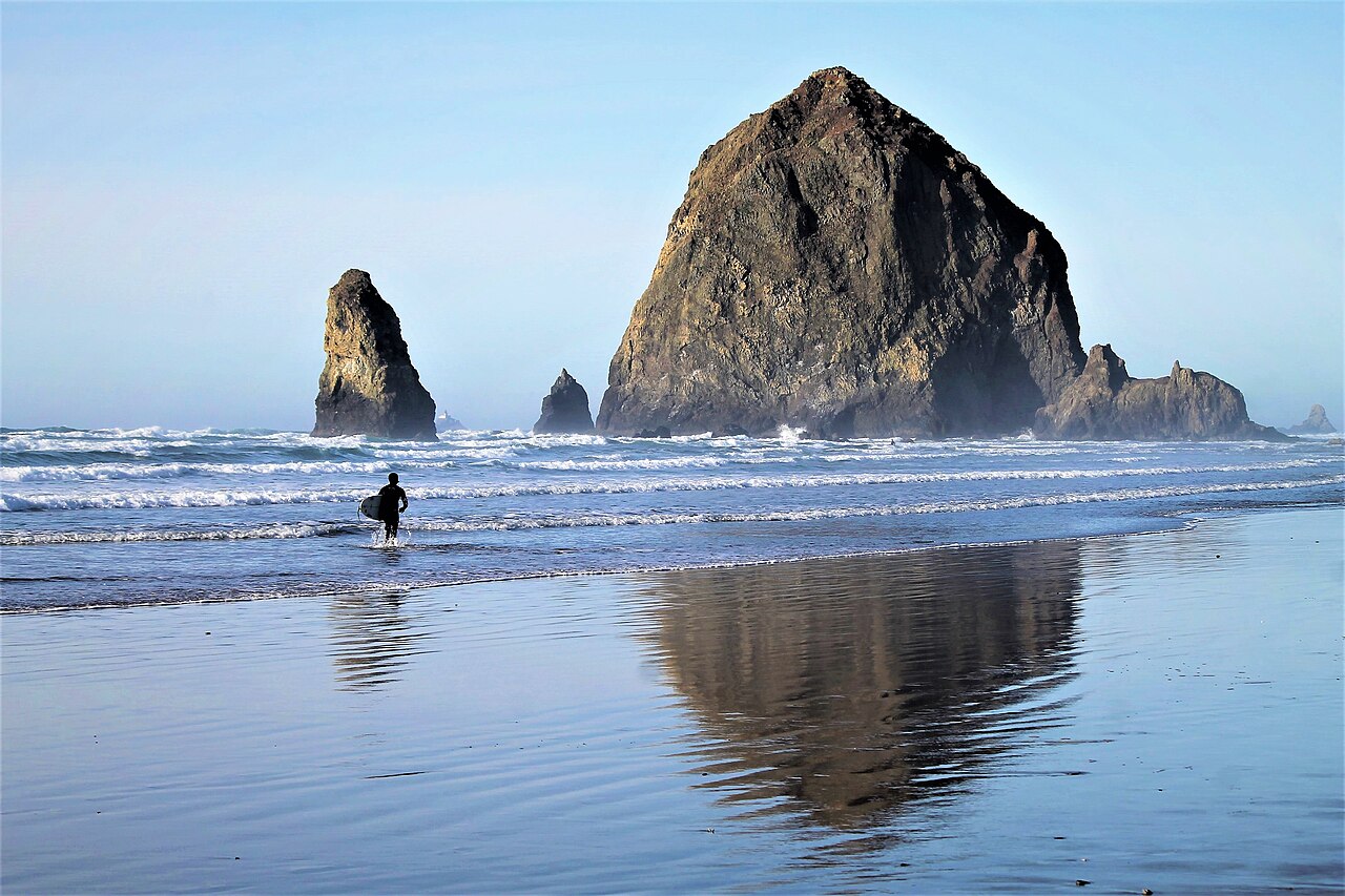 A huge dark sea stack and smaller pointed rocks mirrored in wet sand, with a lone surfer walking toward the waves.