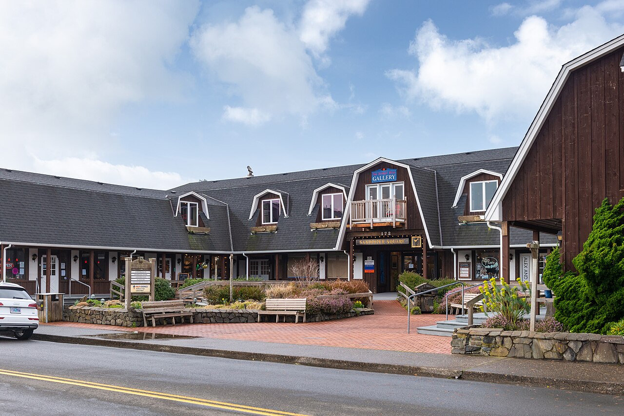 A low row of shops with a grey shingled mansard roof and dormer windows beside a tall brown barn-shaped building.