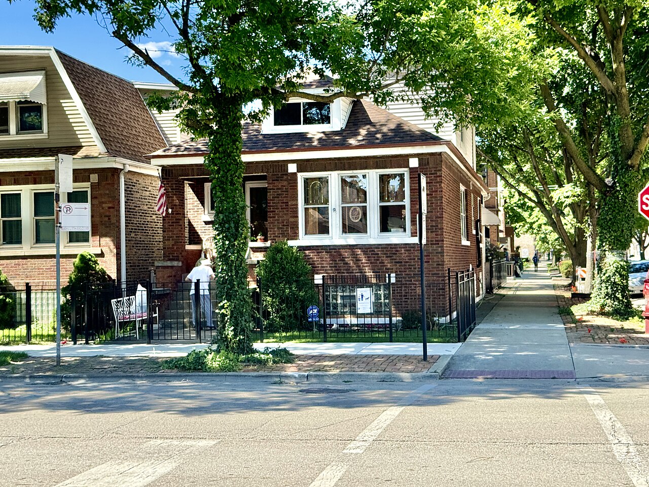 A low brick house with a front porch sits behind an iron fence on a tree-shaded corner, with a pavement running past it.