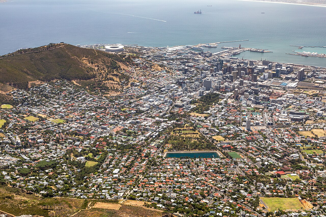 A dense grid of city streets and buildings spreads from the foot of a hill to a harbour and open bay, seen from high above.