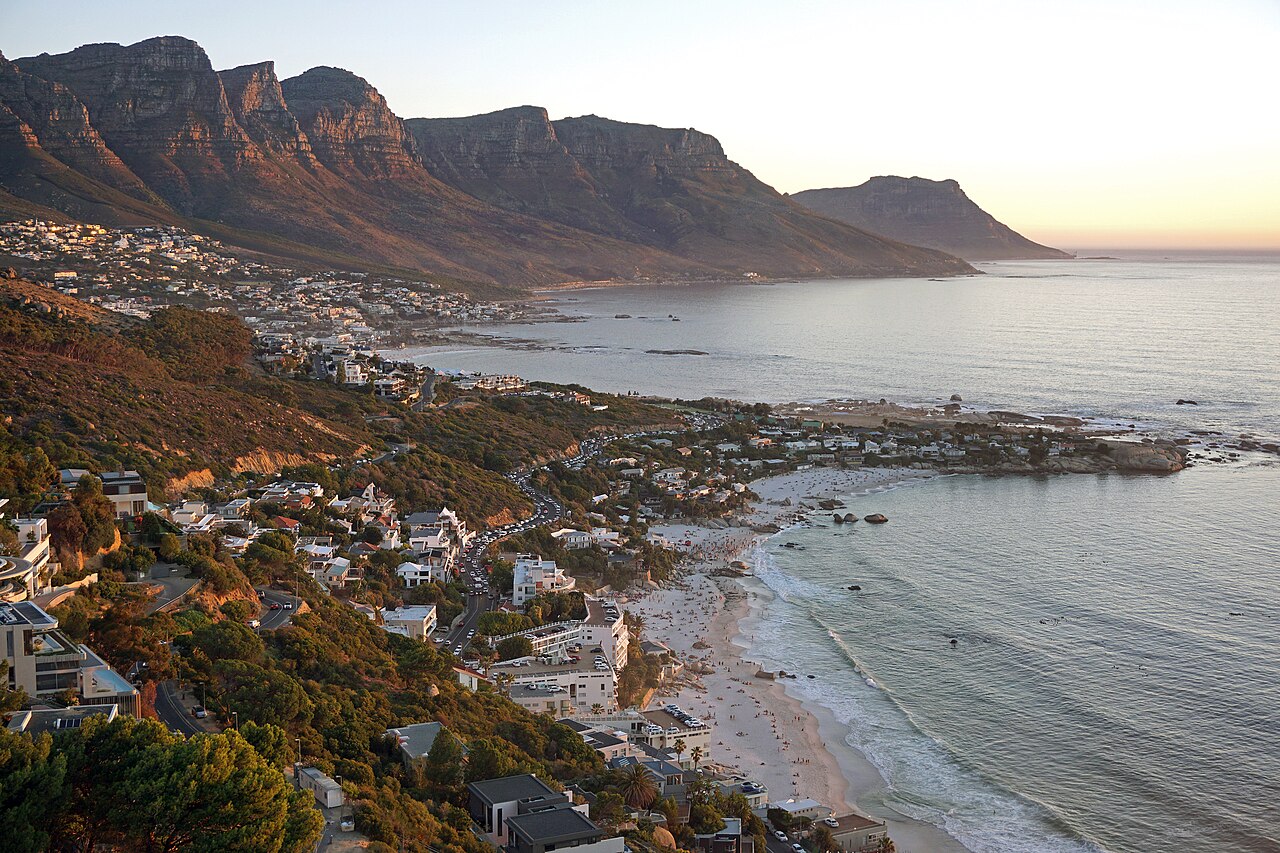 Houses climb a hillside above a calm bay and small beaches, with a row of mountain peaks fading into evening haze.