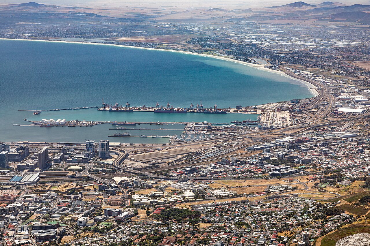 Docks and breakwaters reach into a wide blue bay edged by a long sandy shoreline, with city blocks in the foreground.