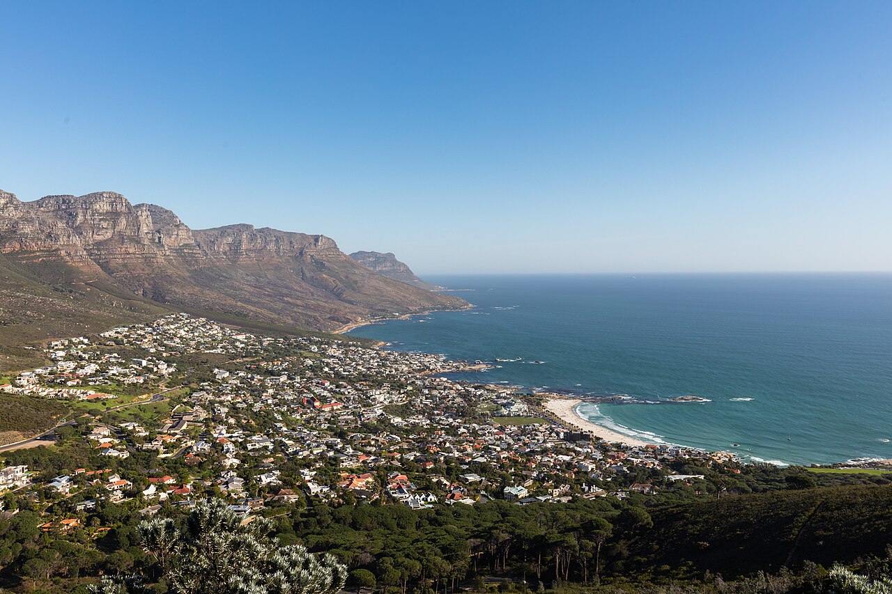A seaside suburb of houses fills the slope between a line of rocky mountain buttresses and a curving bay.