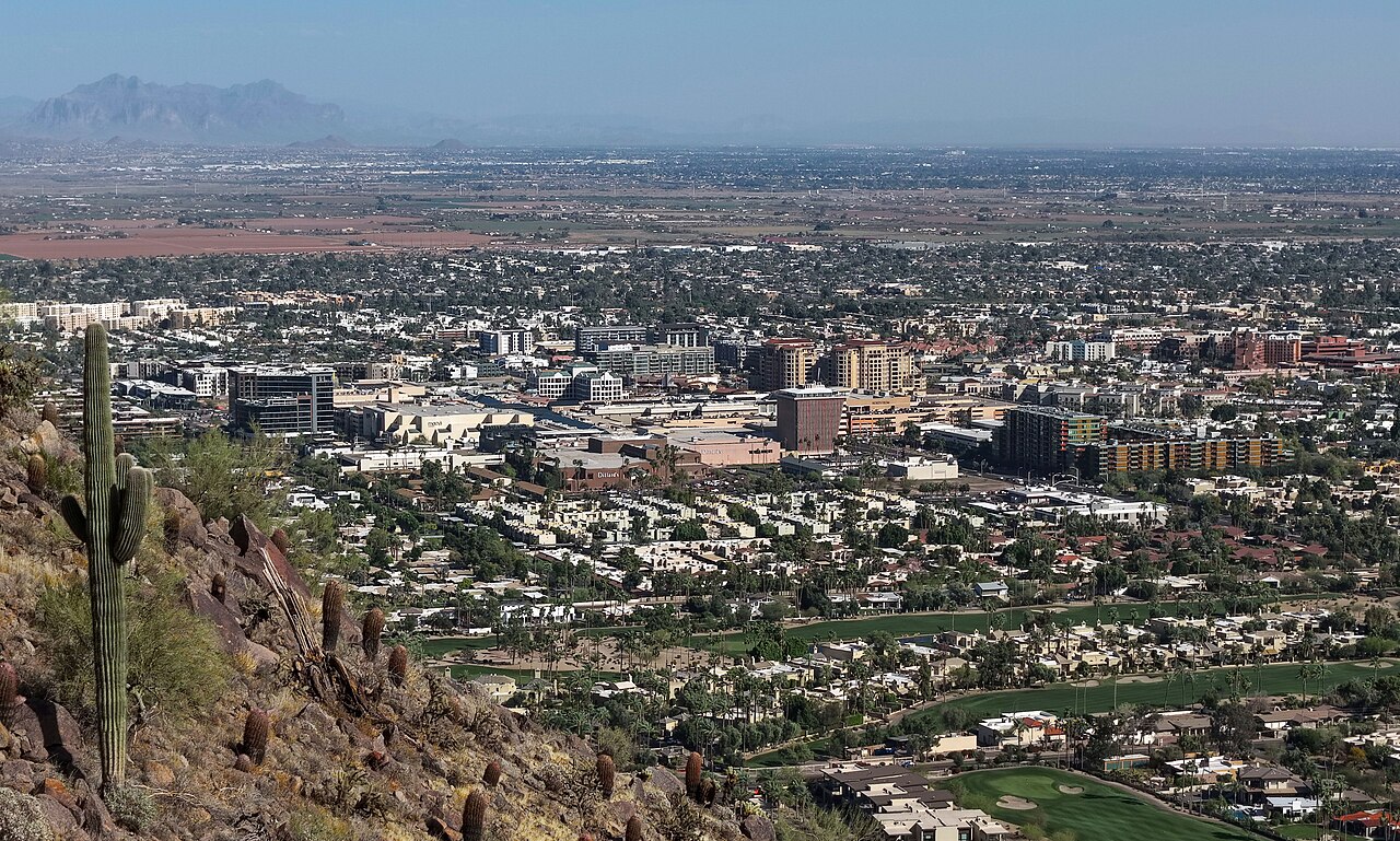 A flat desert city of low buildings, trees and golf greens stretches to distant mountains, seen from a rocky slope.