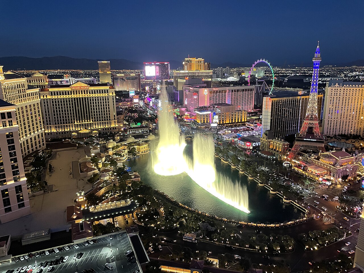 Lit fountains spray from a lake in the middle of a city of bright hotel towers stretching to the horizon at night.