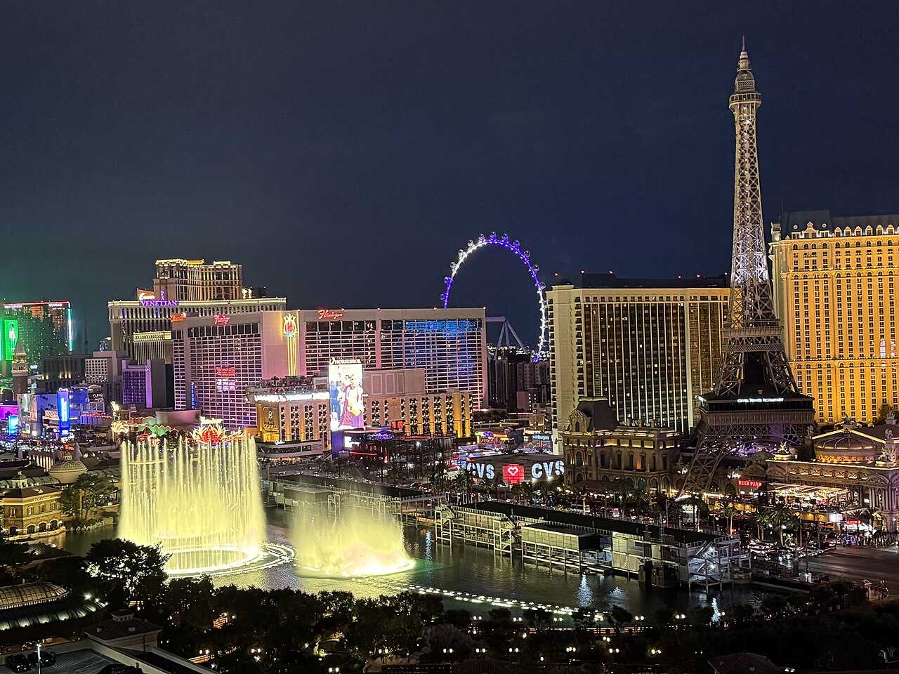 Fountains lit pale yellow rise from a dark lake in front of glowing hotel towers, a replica Eiffel Tower and an observation wheel.