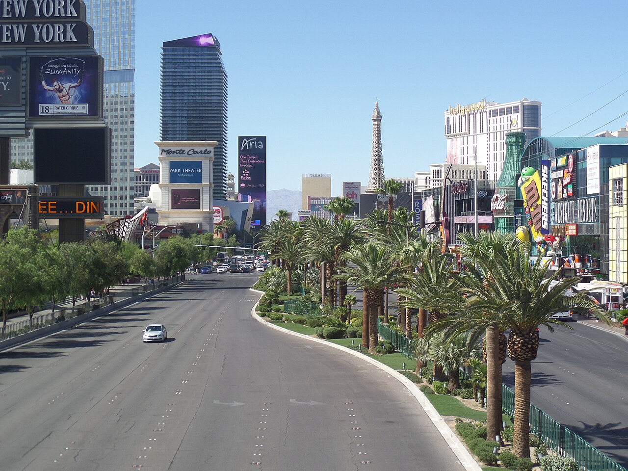 A broad empty boulevard lined with palm trees curves past hotel towers and billboards under a clear sky.