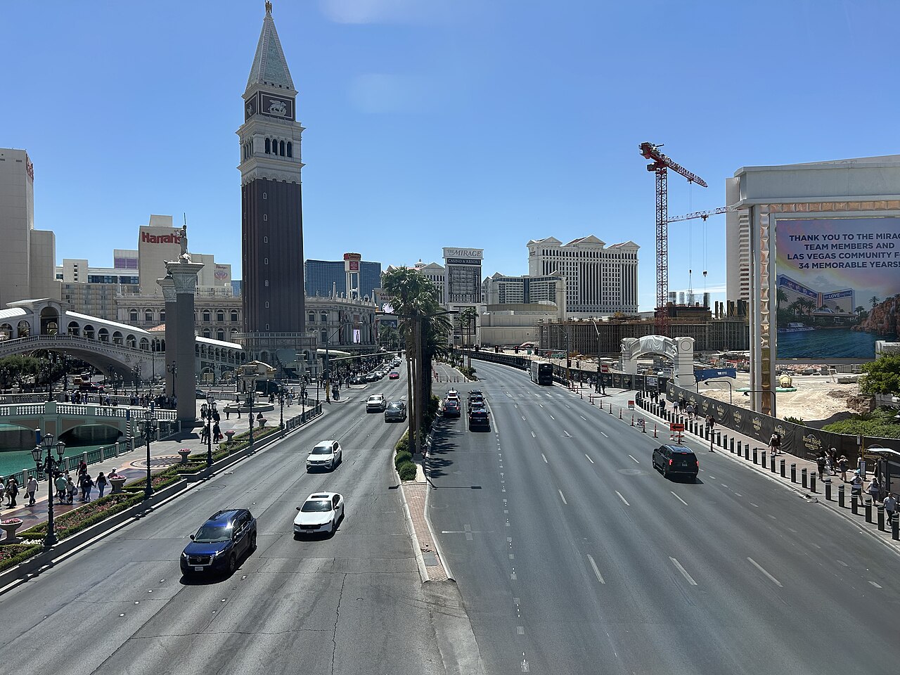A wide multi-lane boulevard with light traffic runs between resort buildings, a tall bell tower and palm trees.