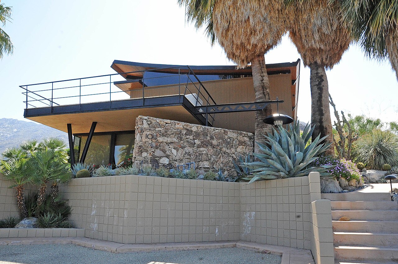 A low modernist house with a curved cantilevered roof and a stone wall sits above a block retaining wall, agaves and palm trunks.