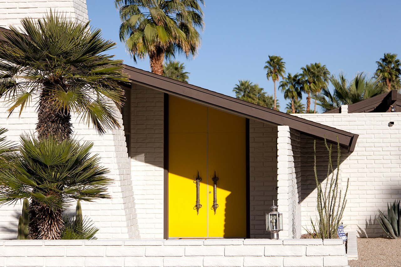 A low white brick house with a sloping dark roof and bright yellow double doors, framed by fan palms.