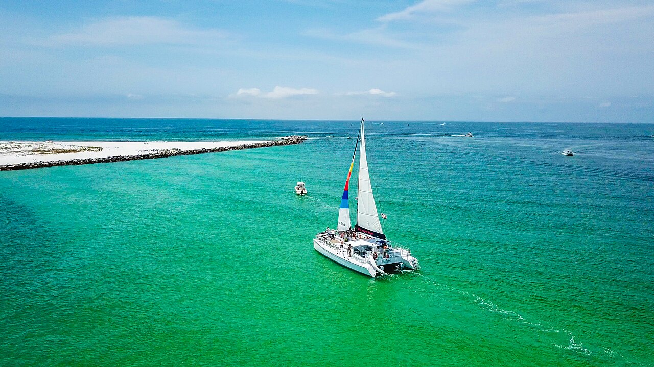 A white catamaran with one sail up crosses bright green shallow water near a rock jetty and a strip of white sand.