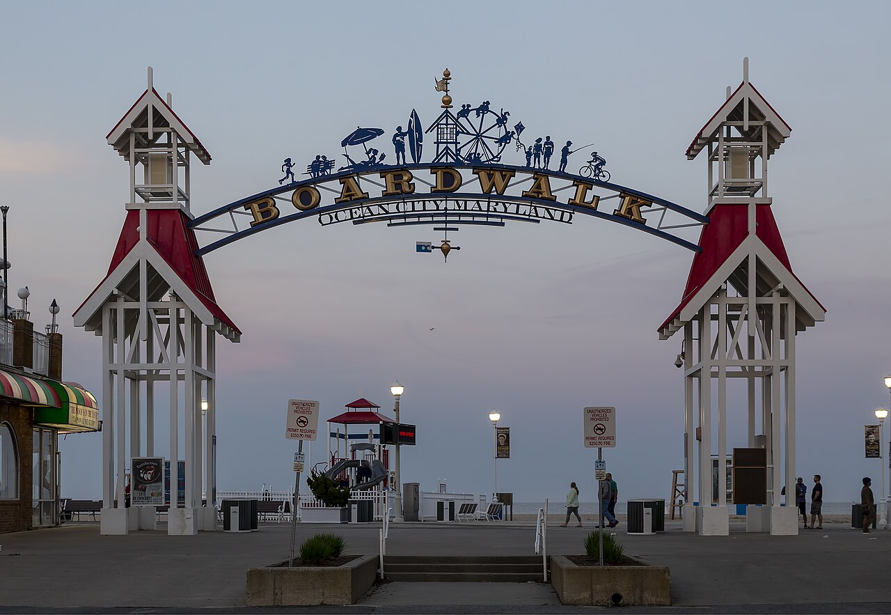 A decorative arch between two small white towers with red roofs spans the entrance to a seaside boardwalk at dusk.