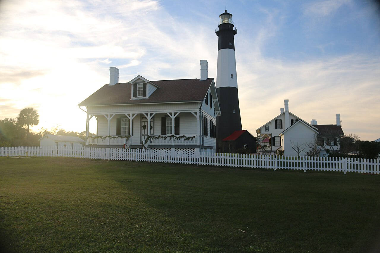 A banded lighthouse and white houses stand in shadow behind a picket fence, lit from behind by a low sun.