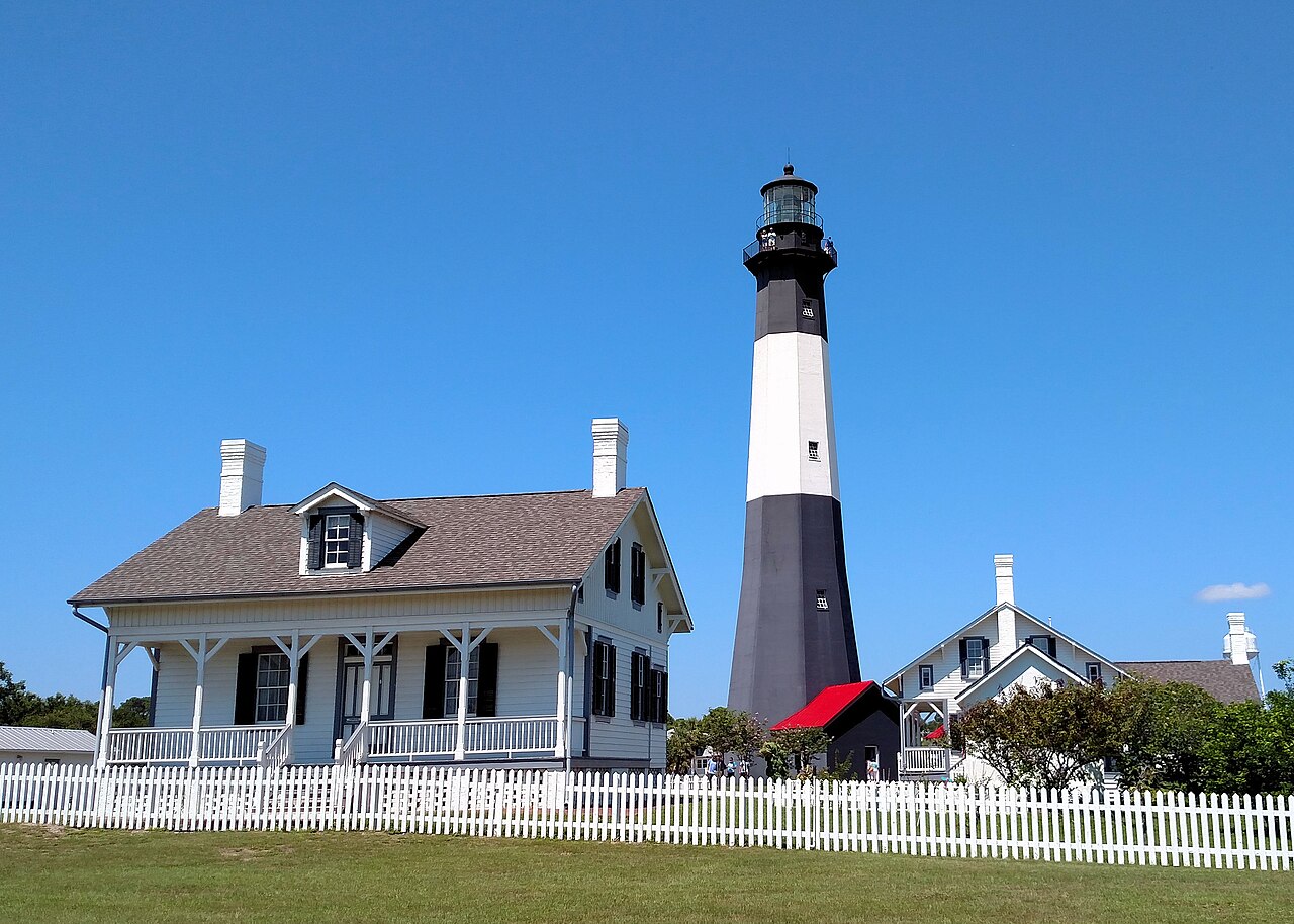 A black and white banded lighthouse stands behind white cottages with grey roofs and a white picket fence.