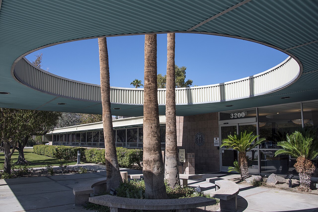 Palm trunks rise through a large circular opening in a flat entrance canopy in front of a low civic building.