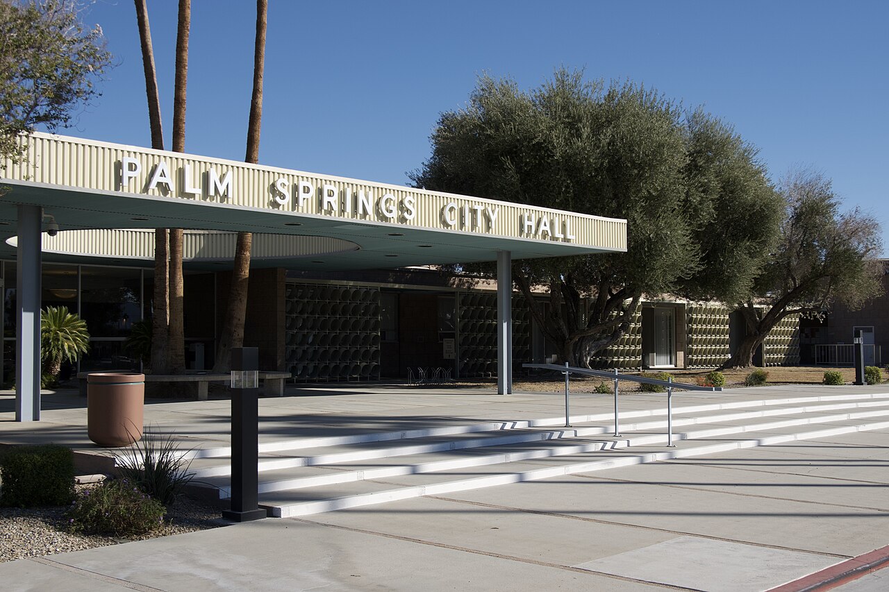 A long flat canopy carrying the building's name in metal letters shades a set of wide steps, with palms and a large tree behind.
