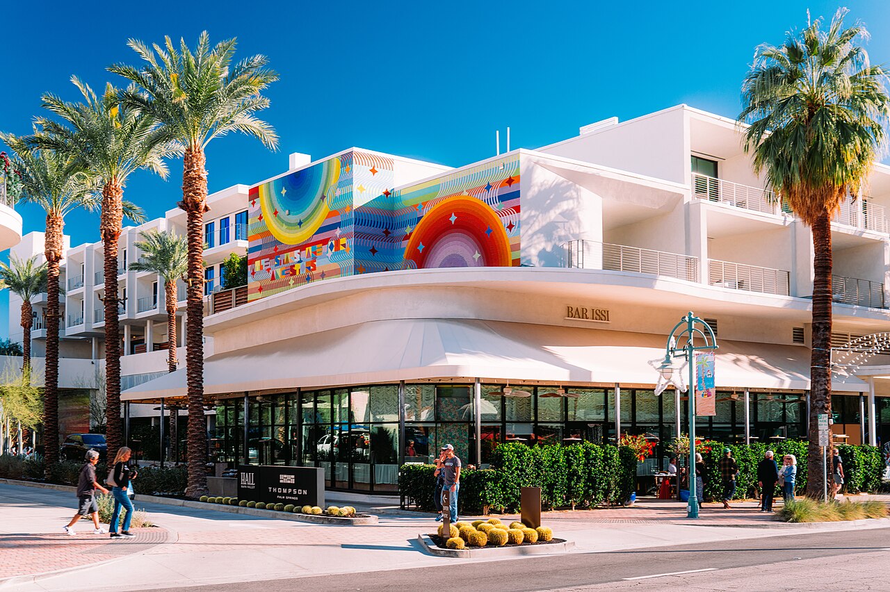 A white three-storey building with a curved awning and a large rainbow mural stands behind tall palm trees on a sunny street.