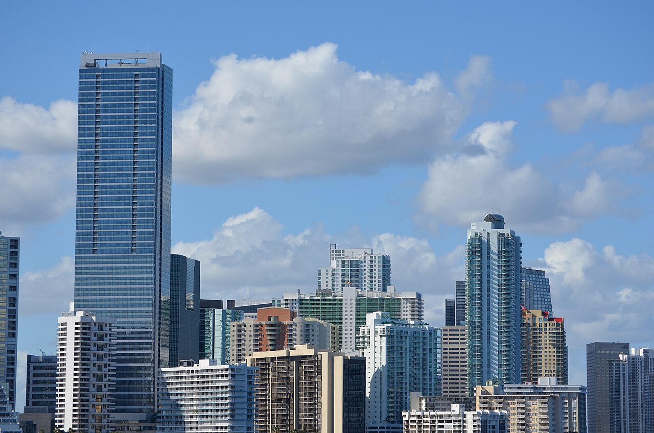 A dense cluster of glass high-rise towers under a blue sky with scattered white clouds.