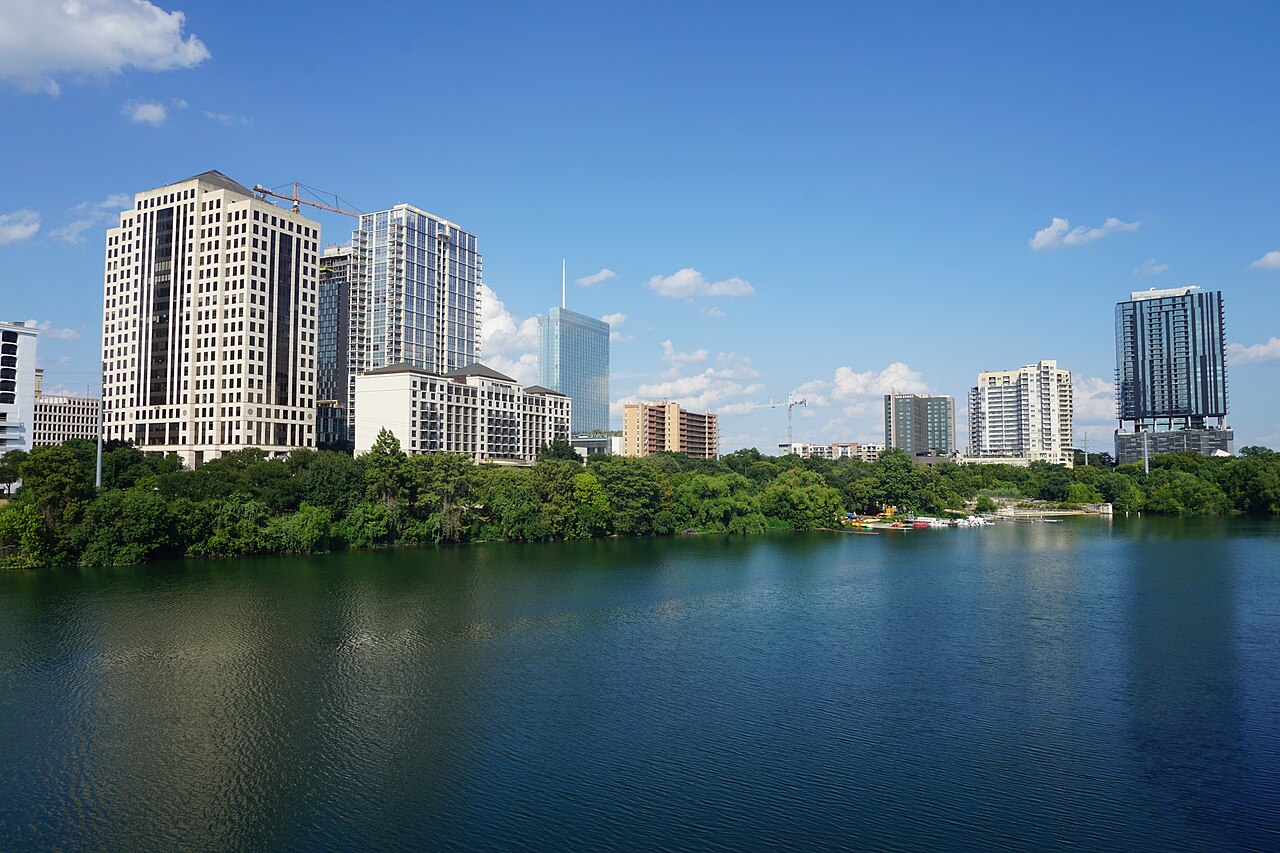 High-rise residential and office towers stand behind a tree-lined shore, with a wide stretch of dark blue water in front.