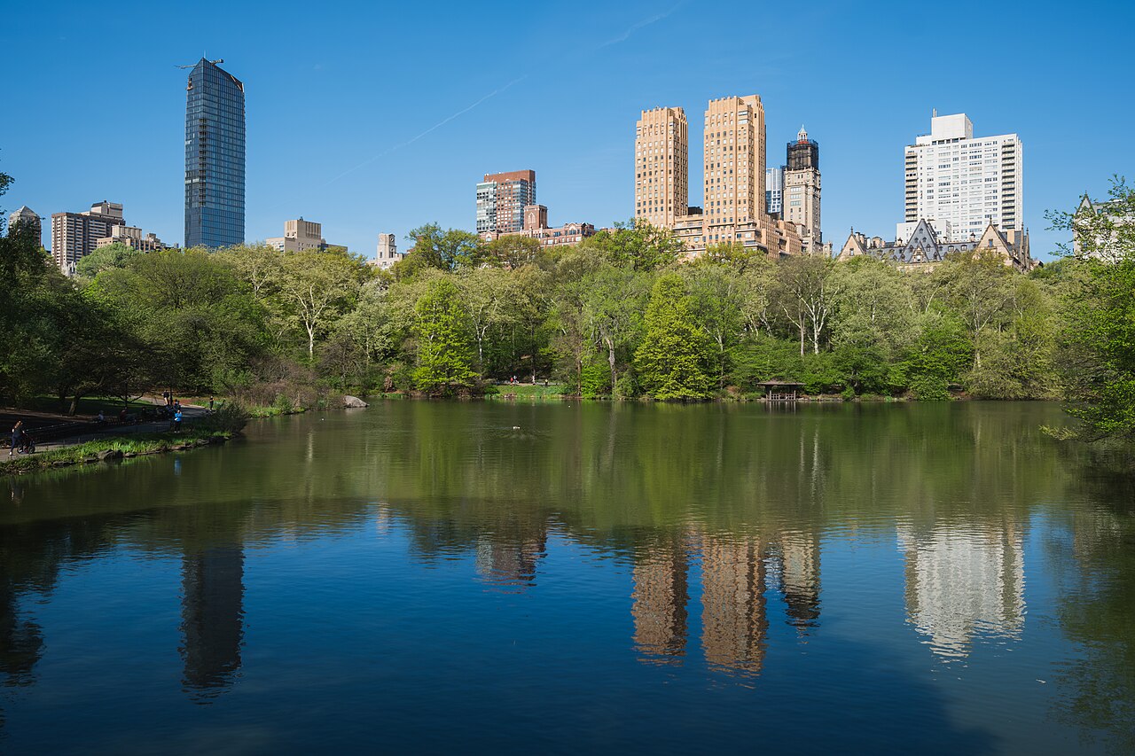 Tall apartment towers rise above a band of green trees and are mirrored in a still lake under a clear blue sky.