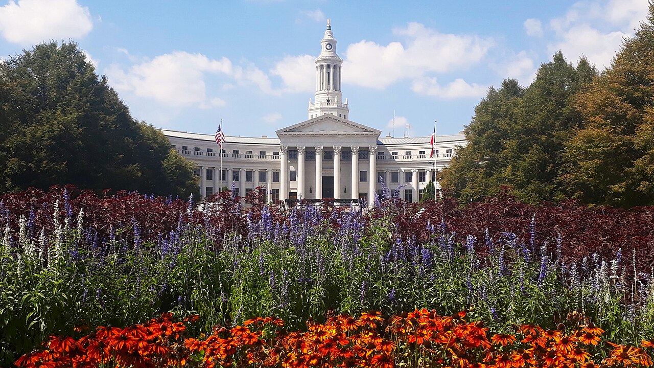 A curved white neoclassical building with a columned entrance and a slender clock tower, behind beds of red and purple flowers.