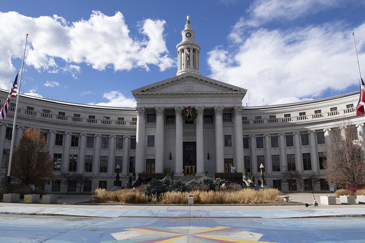A wide grey stone building with curved wings, a columned portico and a central tower, seen across a paved plaza.