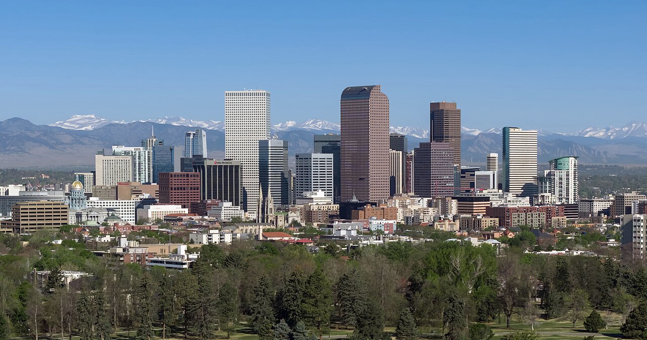 A group of downtown office towers above a belt of trees, with snow-capped mountains along the horizon.