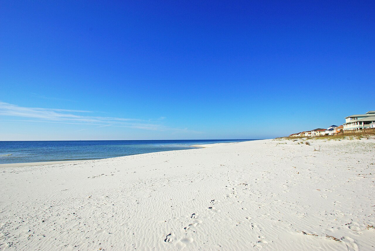A broad, empty stretch of white sand meets calm blue water, with a few low buildings far along the shore.