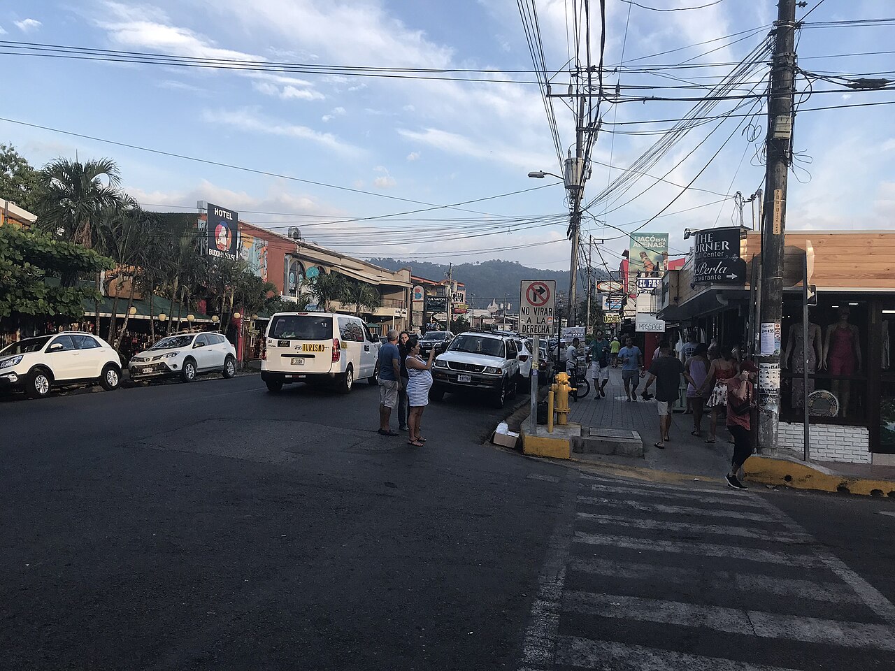 A paved street lined with low shops, parked cars and tangled overhead power lines, with forested hills behind.