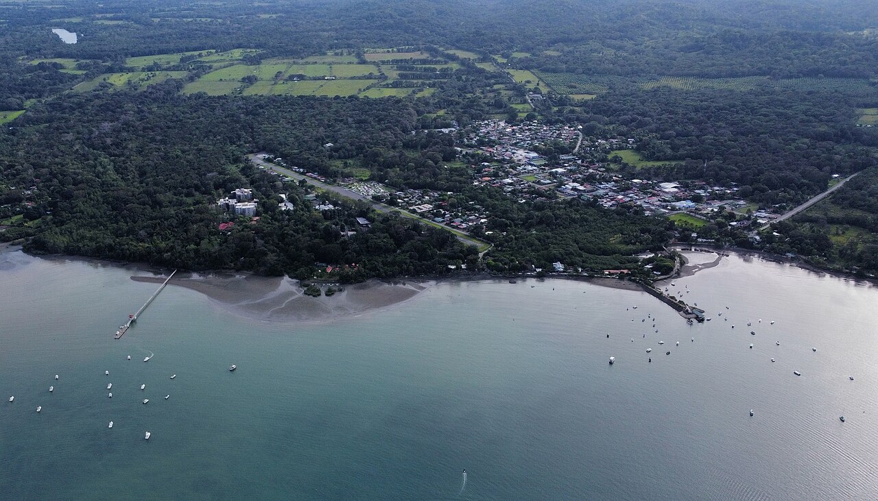An aerial view of a small town among dense green forest beside a calm turquoise bay dotted with moored boats.