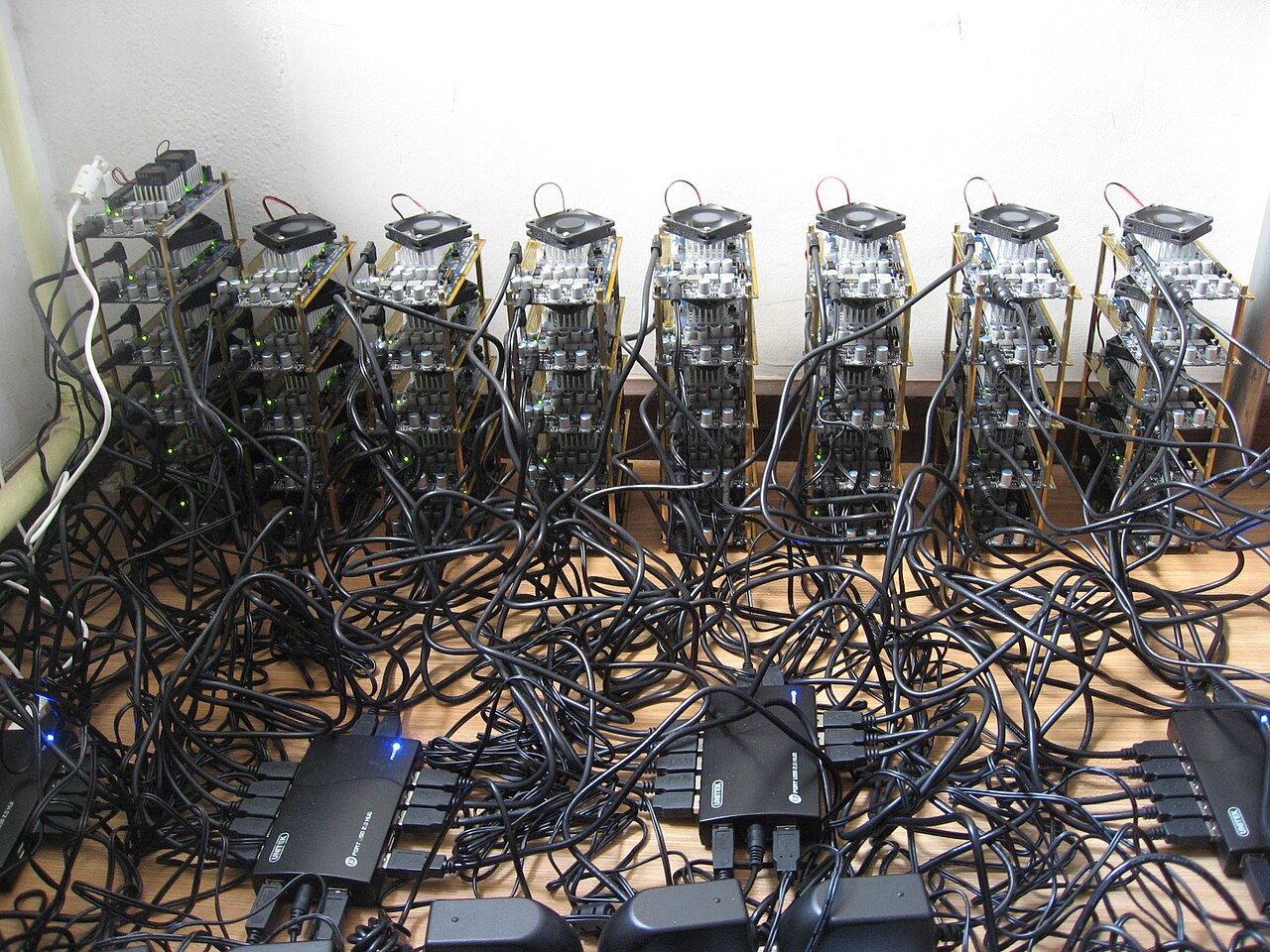 Rows of small circuit boards with cooling fans stand on a table, joined by a thick tangle of black power and data cables.