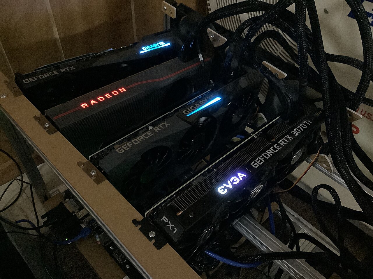 Several graphics cards with glowing logos sit side by side on a metal frame in a dark room, wired together with cables.