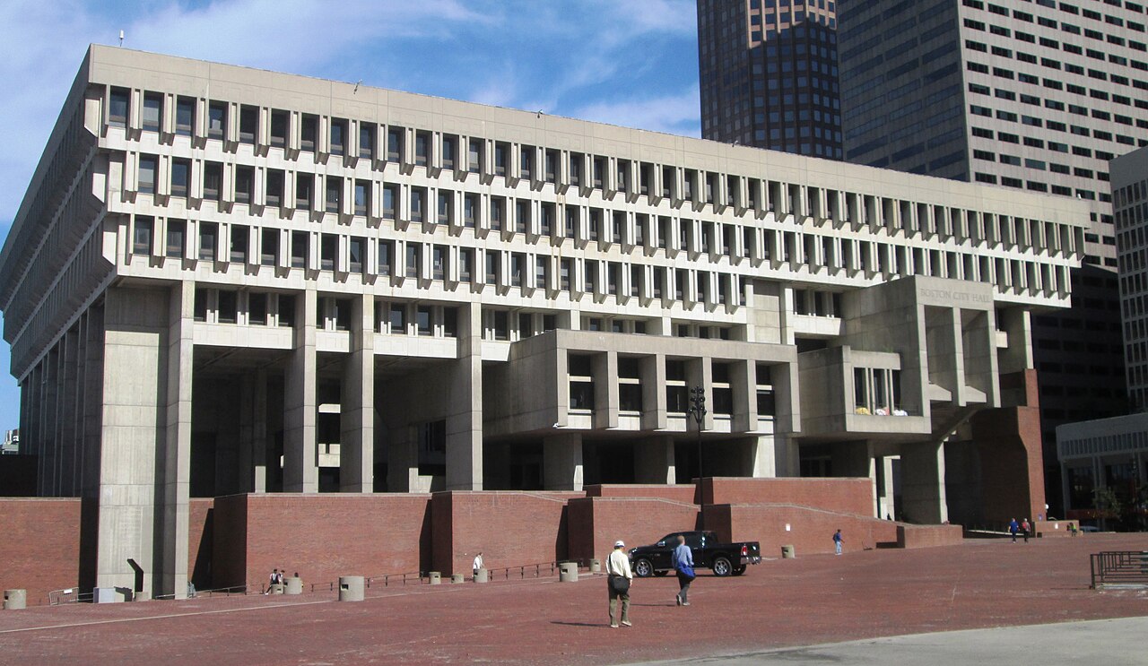 A low-angle view of a concrete building with rows of projecting window bays above a red brick plaza.